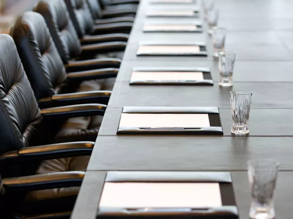 A zoomed picture of the end of a long table with official leather seats lined along it. In equal spacing before the chairs are notepads within a leather case and a pen on the side. In front of each notebook, equally spaced, are crystal water glasses.