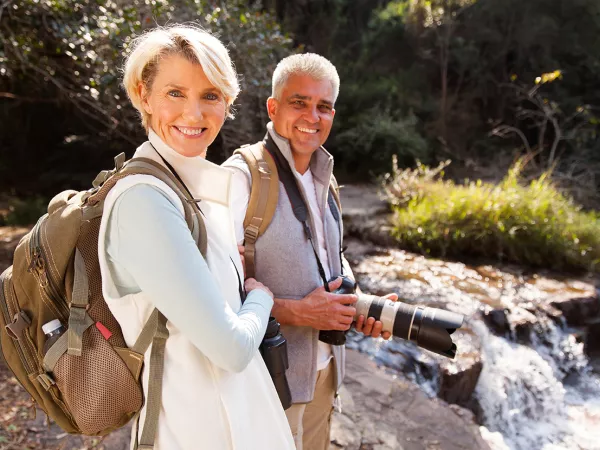 A healthy retired couple hiking along a river with backpacks on and the man is carrying a camera.