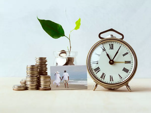 A stack of coins to the left of a glass jar of coins with a growing plant coming out of it, next to an alarm clock and a small picture in the middle front with a couple holding hands and walking on the beach."