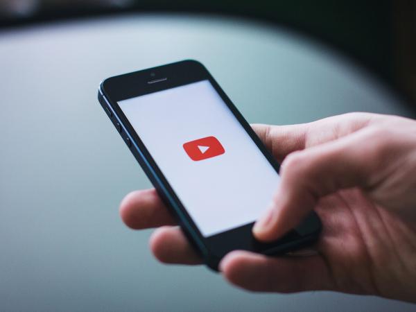 A hand holding a cellphone with a red video play button in the middle of a white background.