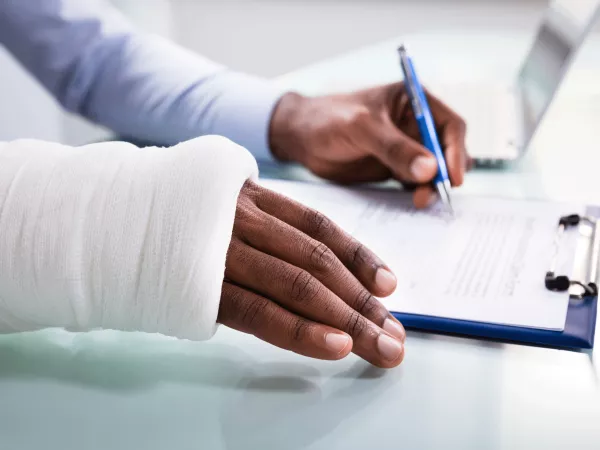 Overhead view of injured man with bandaged hand filling insurance claim form on clipboard.