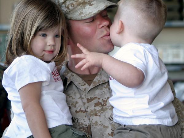 A soldier holding his two small children, giving the youngest a kiss.
