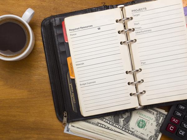 A planner opened to a page with Required Resources:  Person/telephone, Materials, Budget Expenses; and Projects:  Target, Start, Finish, Idea/Summary, Plan. This planner is on top of a calculator and a stack of money sitting next to a cup of coffee.