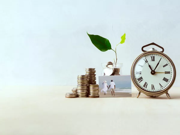 A stack of coins to the left of a glass jar of coins with a growing plant coming out of it, next to an alarm clock and a small picture in the middle front with a couple holding hands and walking on the beach."