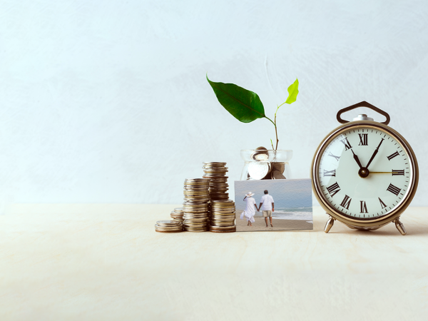 A stack of coins to the left of a glass jar of coins with a growing plant coming out of it, next to an alarm clock and a small picture in the middle front with a couple holding hands and walking on the beach."