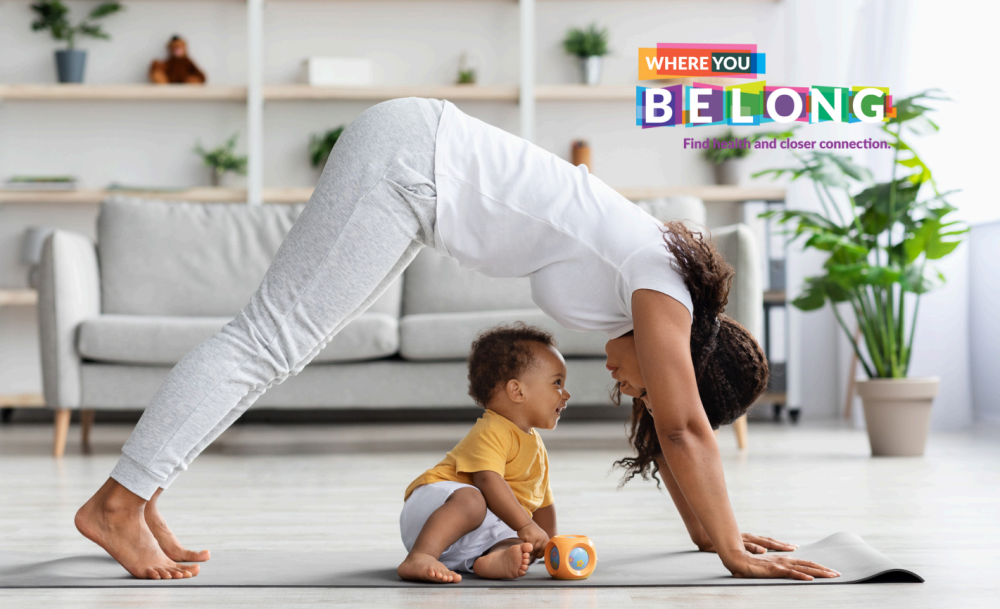 Mother stretching in downward dog pose, with child sitting on the mat below the mother's core