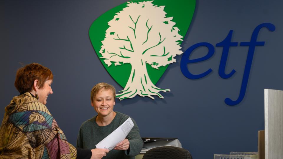 ETF front desk person assisting someone by showing her a piece of paper. The ETF logo is prominent in the background wall.
