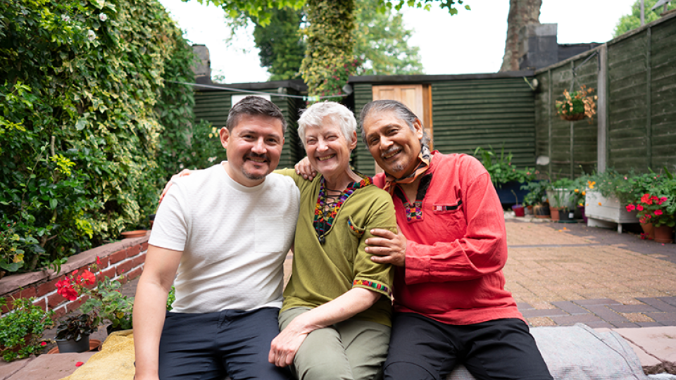 Three people smiling and sitting on a bench in a high-fenced backyard