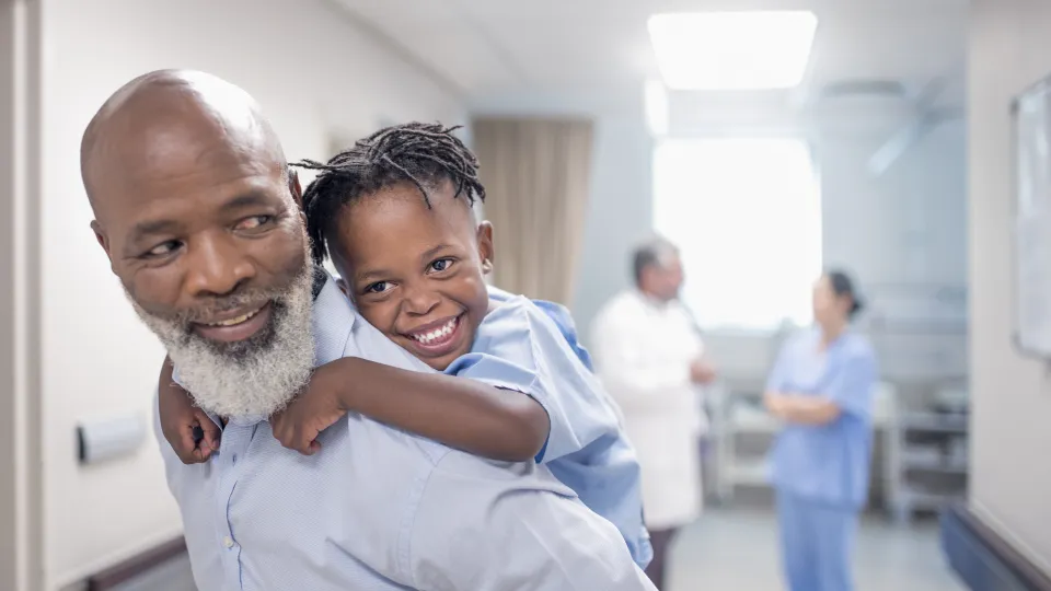 A parent piggybacks his child, with two healthcare workers in the background.