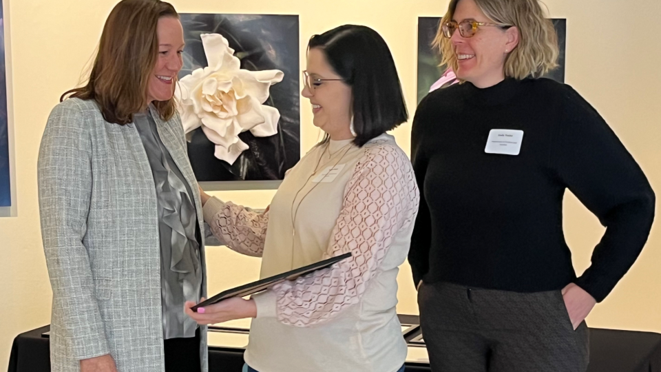 Two women from the Department of Children and Families receive the award certificate from Jen Flogel of the Department of Administration. All three women are standing, with photos of flowers behind them.