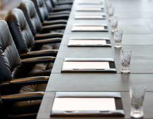 A zoomed picture of the end of a long table with official leather seats lined along it. In equal spacing before the chairs are notepads within a leather case and a pen on the side. In front of each notebook, equally spaced, are crystal water glasses.