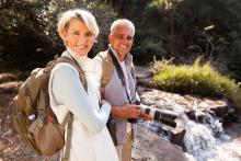 A healthy retired couple hiking along a river with backpacks on and the man is carrying a camera.