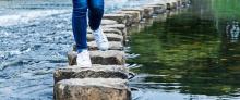 Person stepping on a bridge of stones to cross water.