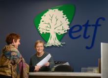 ETF front desk person assisting someone by showing her a piece of paper. The ETF logo is prominent in the background wall.