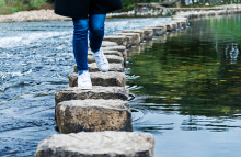 Pair of legs in blue jeans with white sneakers walking through steppingstones in a body of water