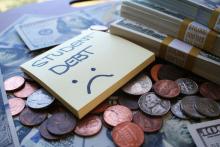 A sticky note sitting over money with the words, "STUDENT DEBT" and an unhappy face.