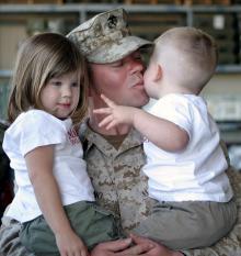 A soldier holding his two small children, giving the youngest a kiss.