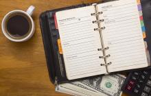 A planner opened to a page with Required Resources:  Person/telephone, Materials, Budget Expenses; and Projects:  Target, Start, Finish, Idea/Summary, Plan. This planner is on top of a calculator and a stack of money sitting next to a cup of coffee.
