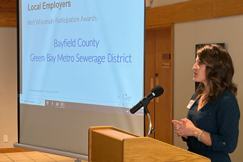 Well Wisconsin's Molly Dunks stands behind a lectern beside a presentation screen with the texts: Local Employers, Well Wisconsin Participation Awards, Bayfield County, Green Bay Metro Sewerage District