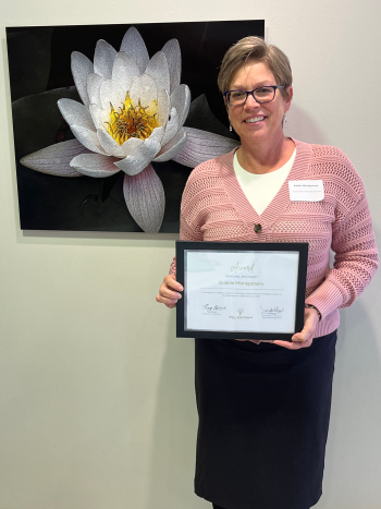 Bobbie Montgomery shows her framed award certificate while standing in front of a large photo of a white flower